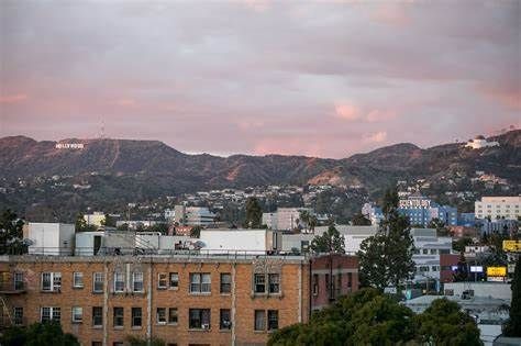 A multi-story brick apartment building in the foreground, with the Hollywood Sign visible on hills under a pink sky.