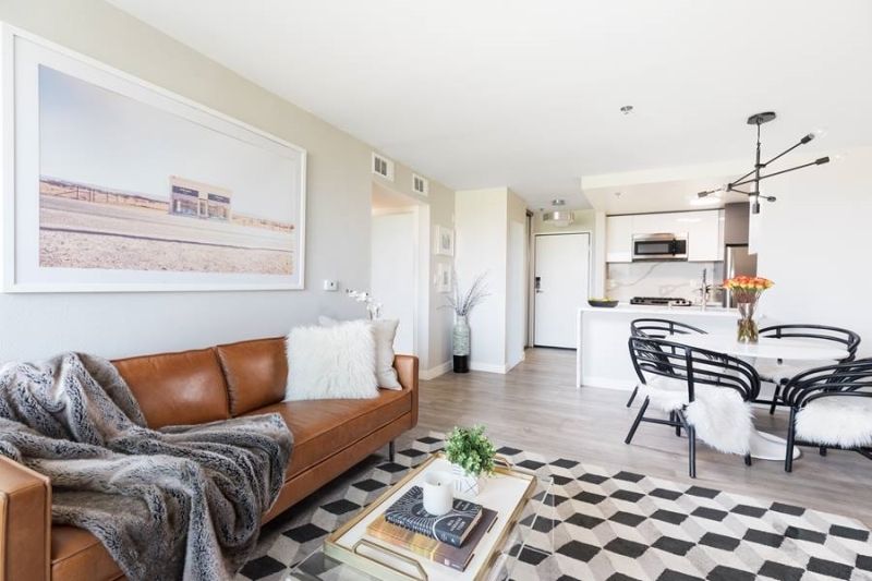A bright living and dining room with a brown leather sofa, white accents, a geometric rug, and an open-concept kitchen.