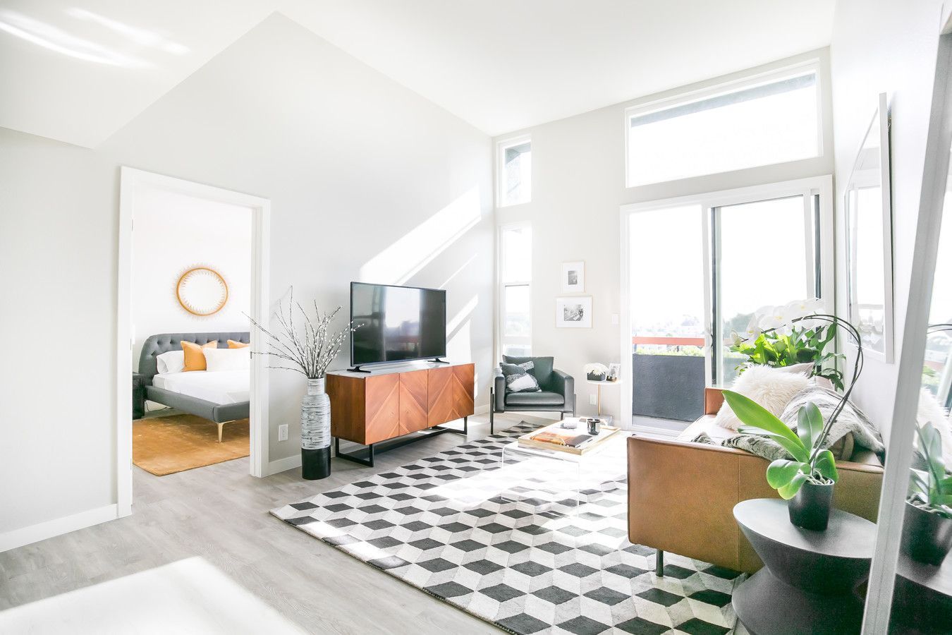 A bright living room with white walls, a geometric rug, a leather sofa, and a wood media console, open to a bedroom.