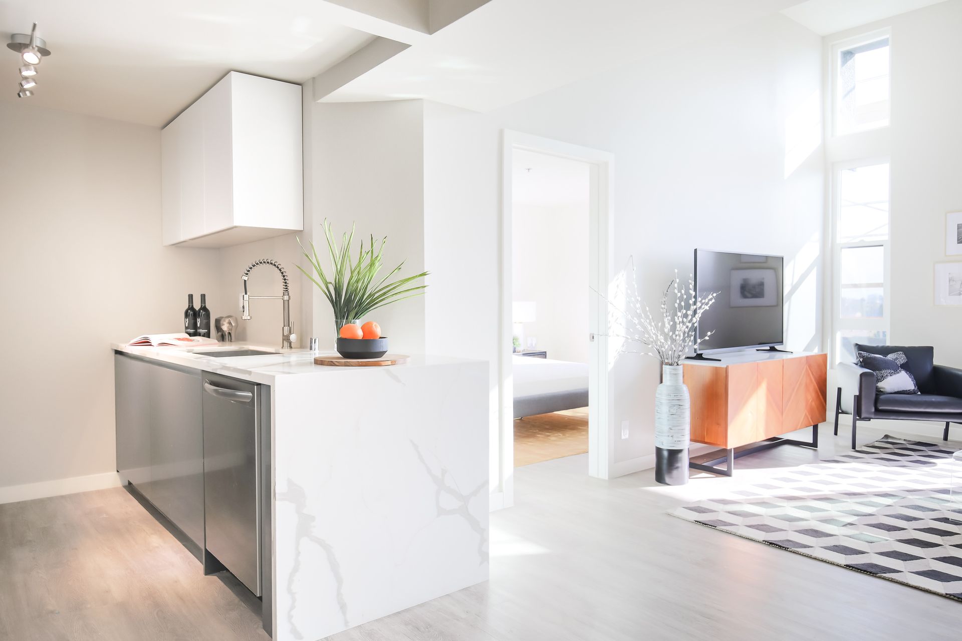 Modern apartment interior with a minimalist kitchen featuring white stone counters, a bedroom doorway, and living area.