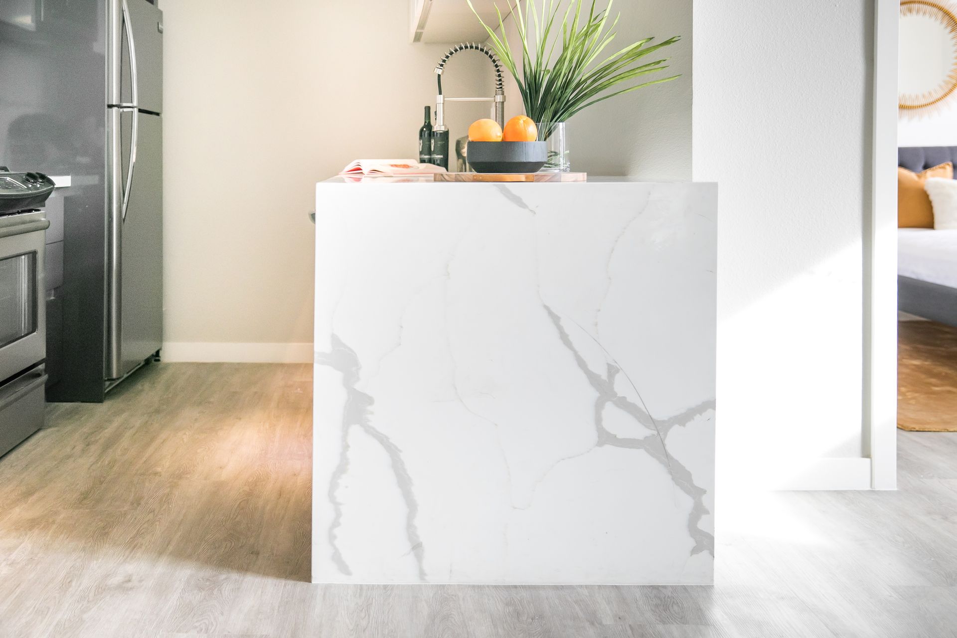 A white, marble-patterned kitchen island stands on a light wood floor in a modern kitchen with a refrigerator nearby.