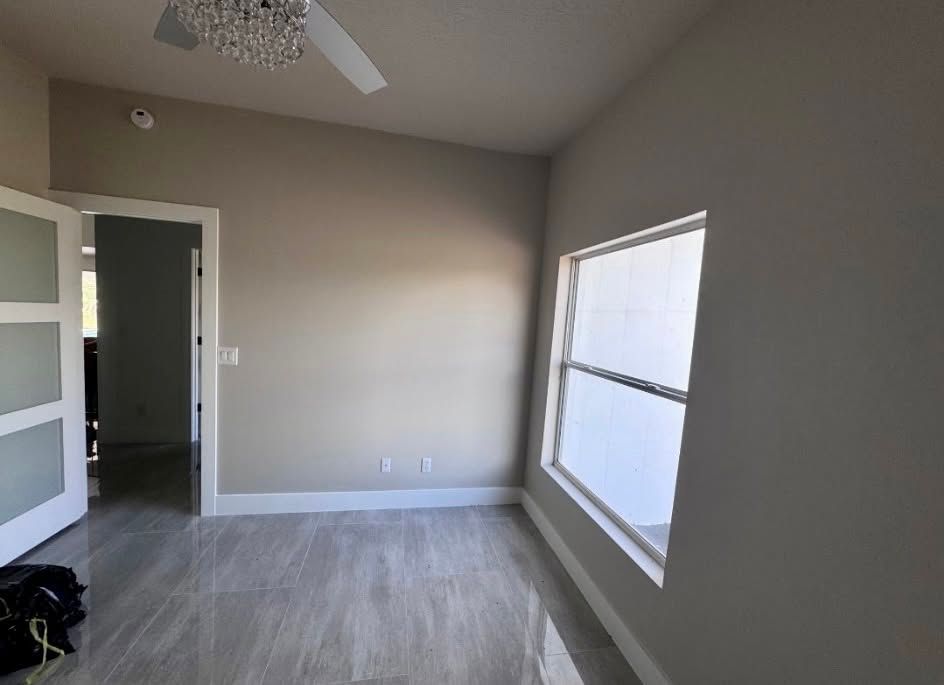 Empty beige bedroom with gray flooring, ceiling fan, open doorway, and large window with white trim