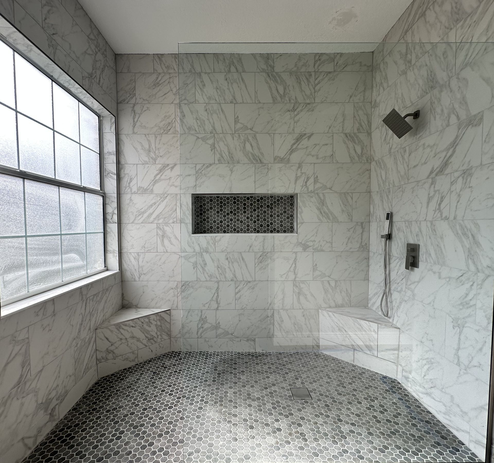 Marble-tiled shower with large window, bench, wall niche, and mosaic pebble floor.