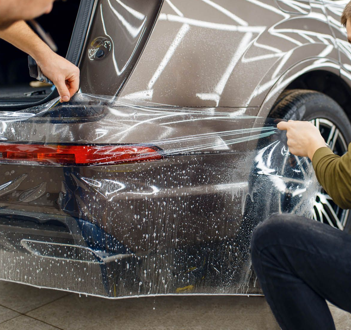 Two people wrapping clear protective film around a gray car’s rear bumper in a garage