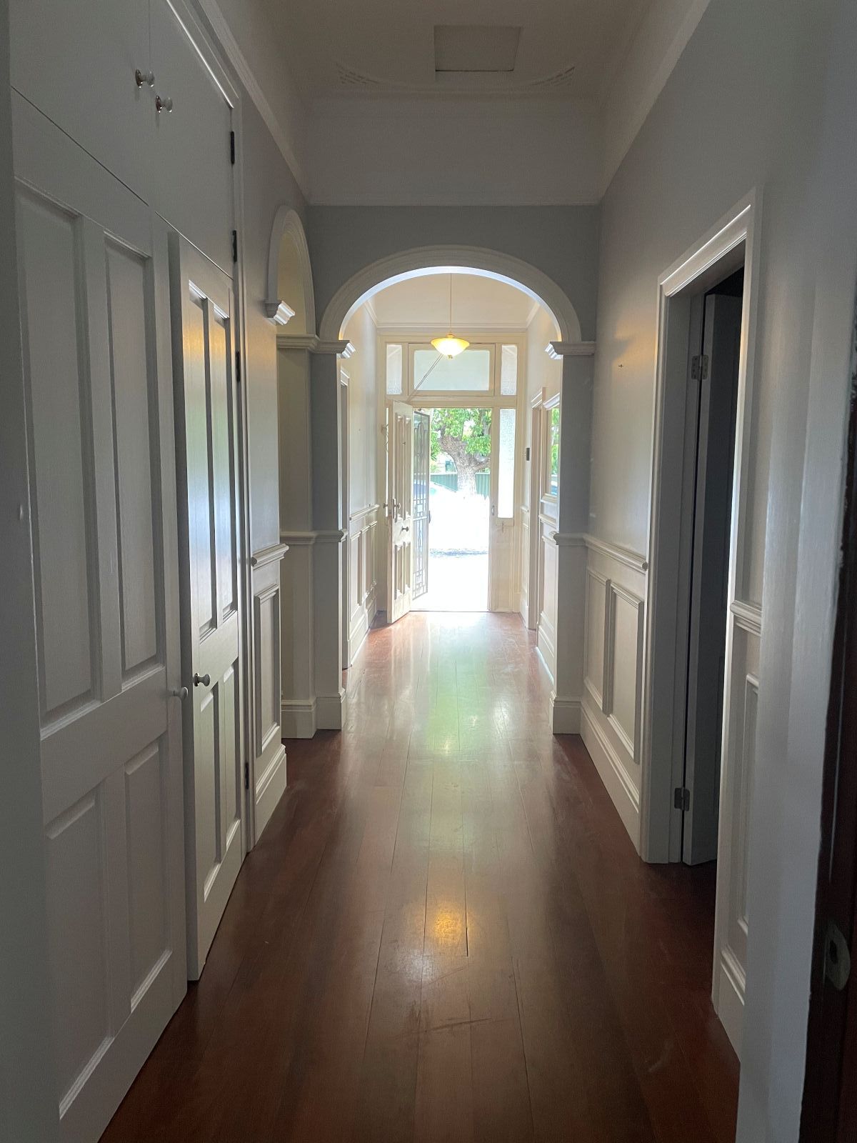 Long hallway with wood floor, white walls, arched doorways, and sunlight at the end.
