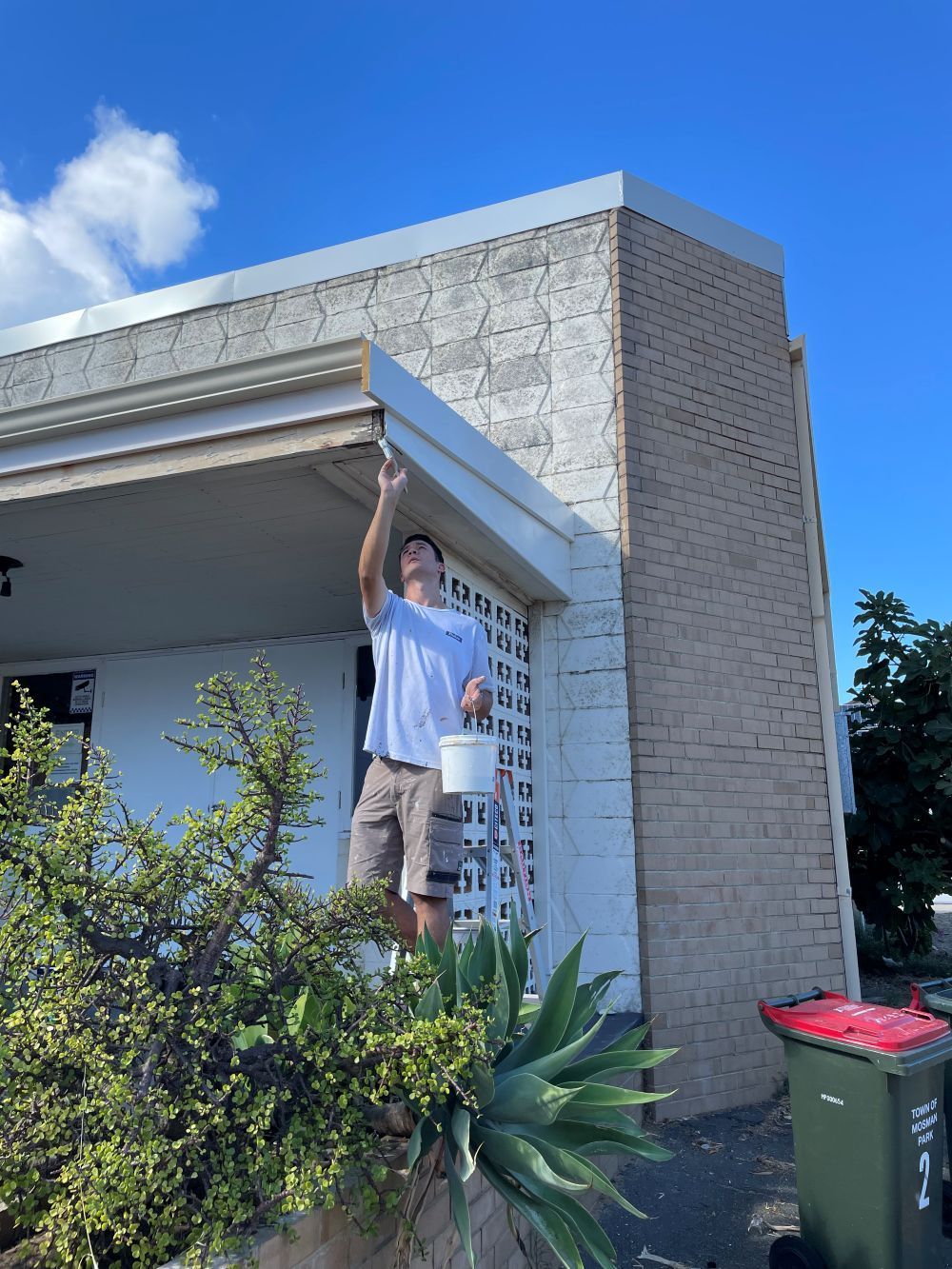 Man on a ladder painting the trim of a two-story brick building on a sunny day.