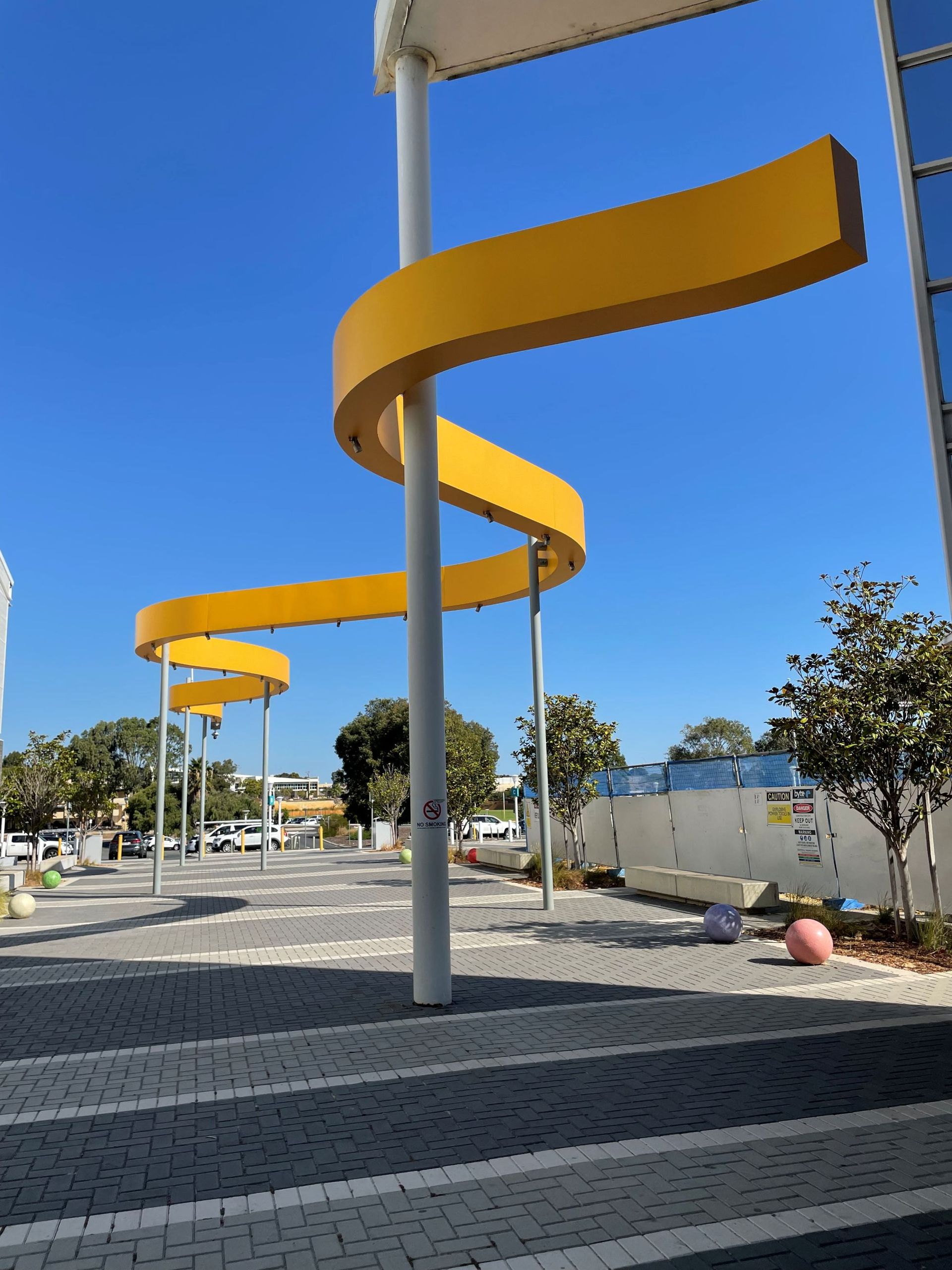 Fresh painted yellow, undulating, art installation with white supports against a blue sky in an outdoor plaza.