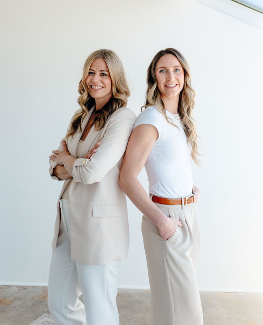 Two women in light neutral outfits standing back-to-back in a bright, minimalist room