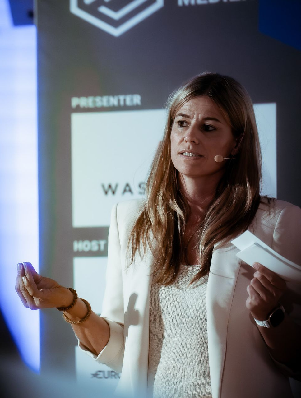 Speaker presenting on stage with microphone, white blazer, and event backdrop behind her