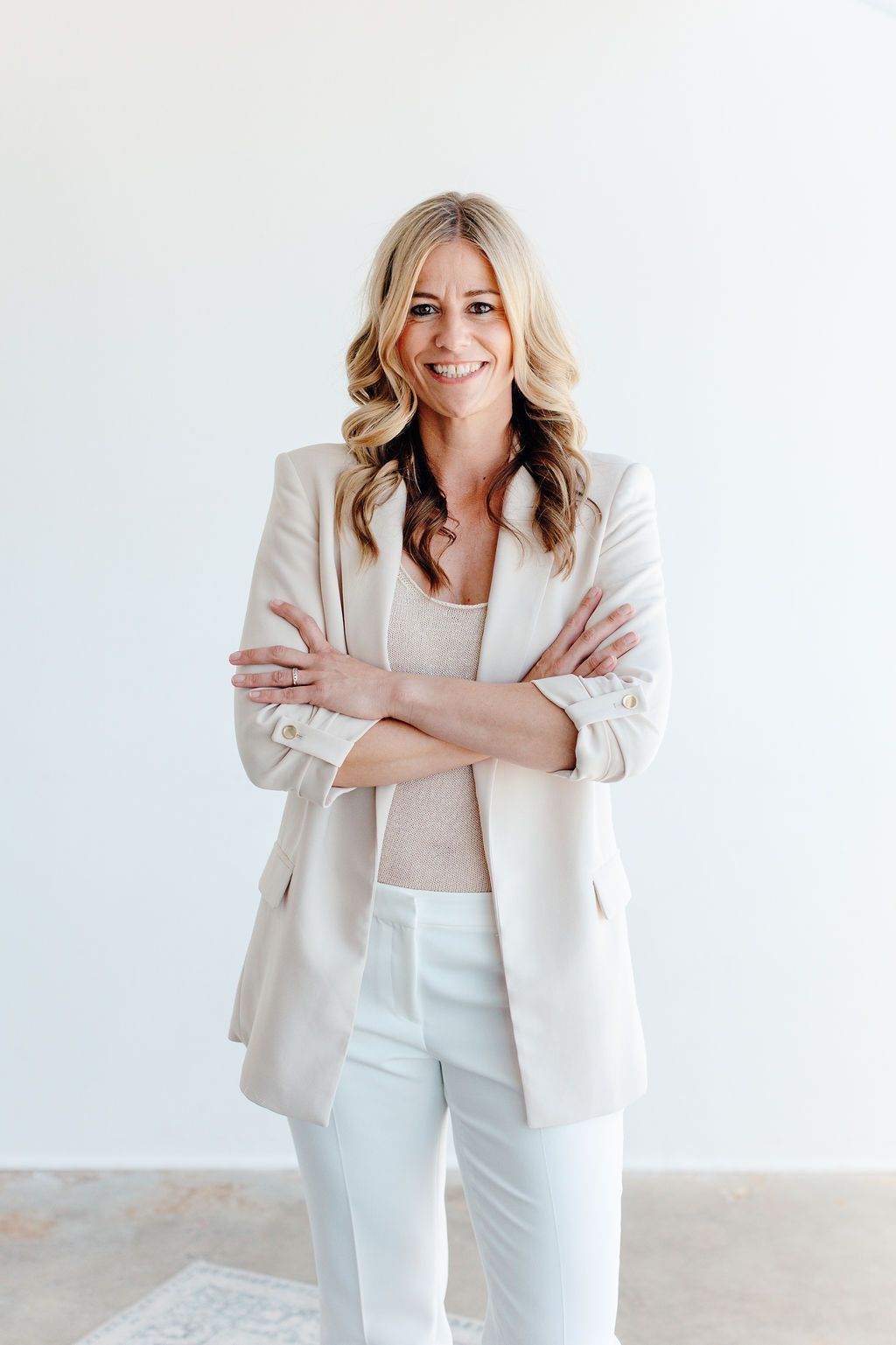 Smiling woman in a white blazer and pants standing with arms crossed against a plain light background