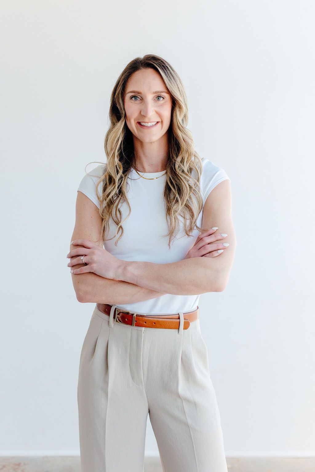 Smiling woman in a white shirt and beige pants stands with arms crossed against a plain light wall