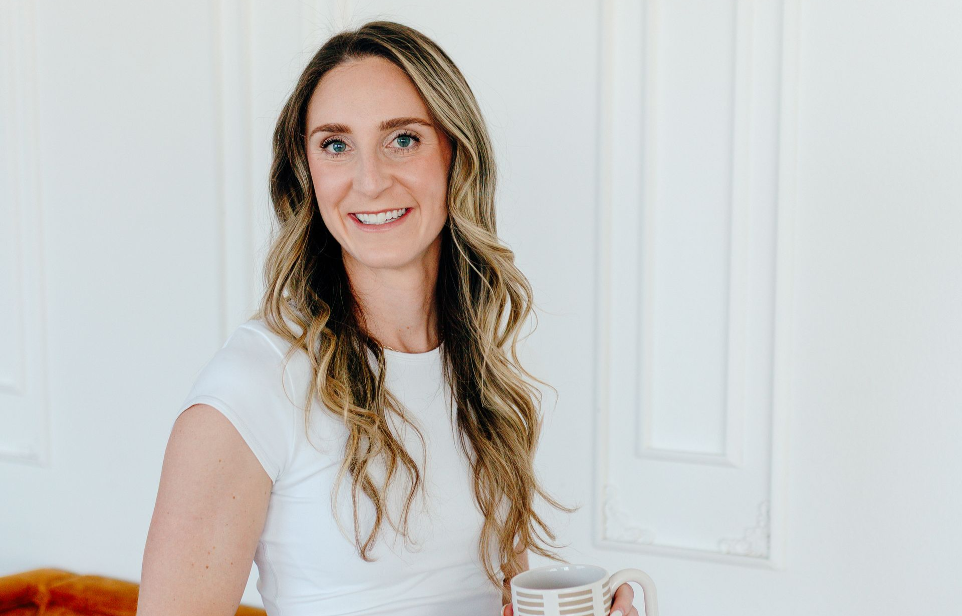 Woman smiling in a white room, wearing a white top and holding a mug.