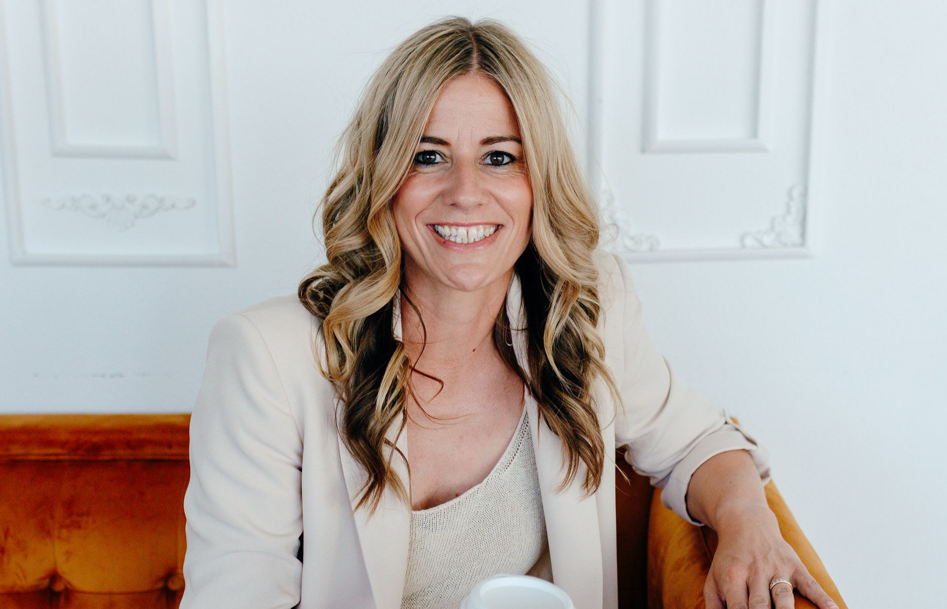 Smiling woman seated on a wooden bench, wearing a light blazer and lace top against a white paneled wall.