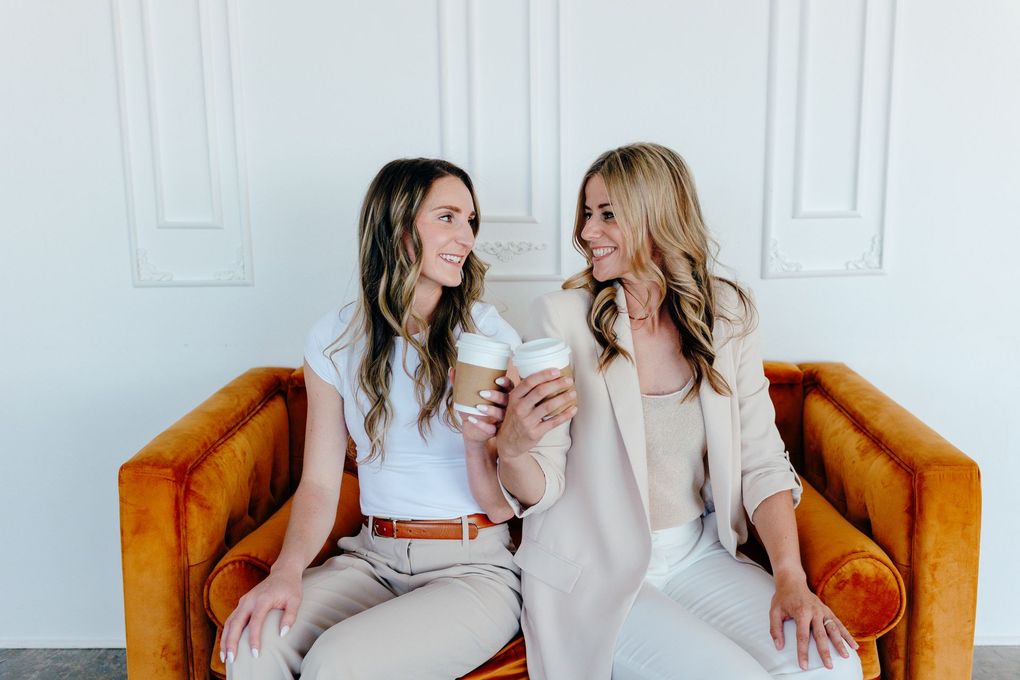 Two people sitting on an orange couch, smiling at each other and holding drinks in a bright room.