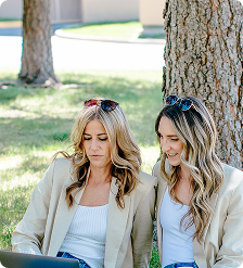 Two women seated outdoors under trees, looking down at a laptop together