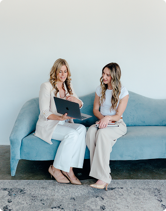 Two women sit on a light blue sofa, looking at a laptop in a bright, minimal room.
