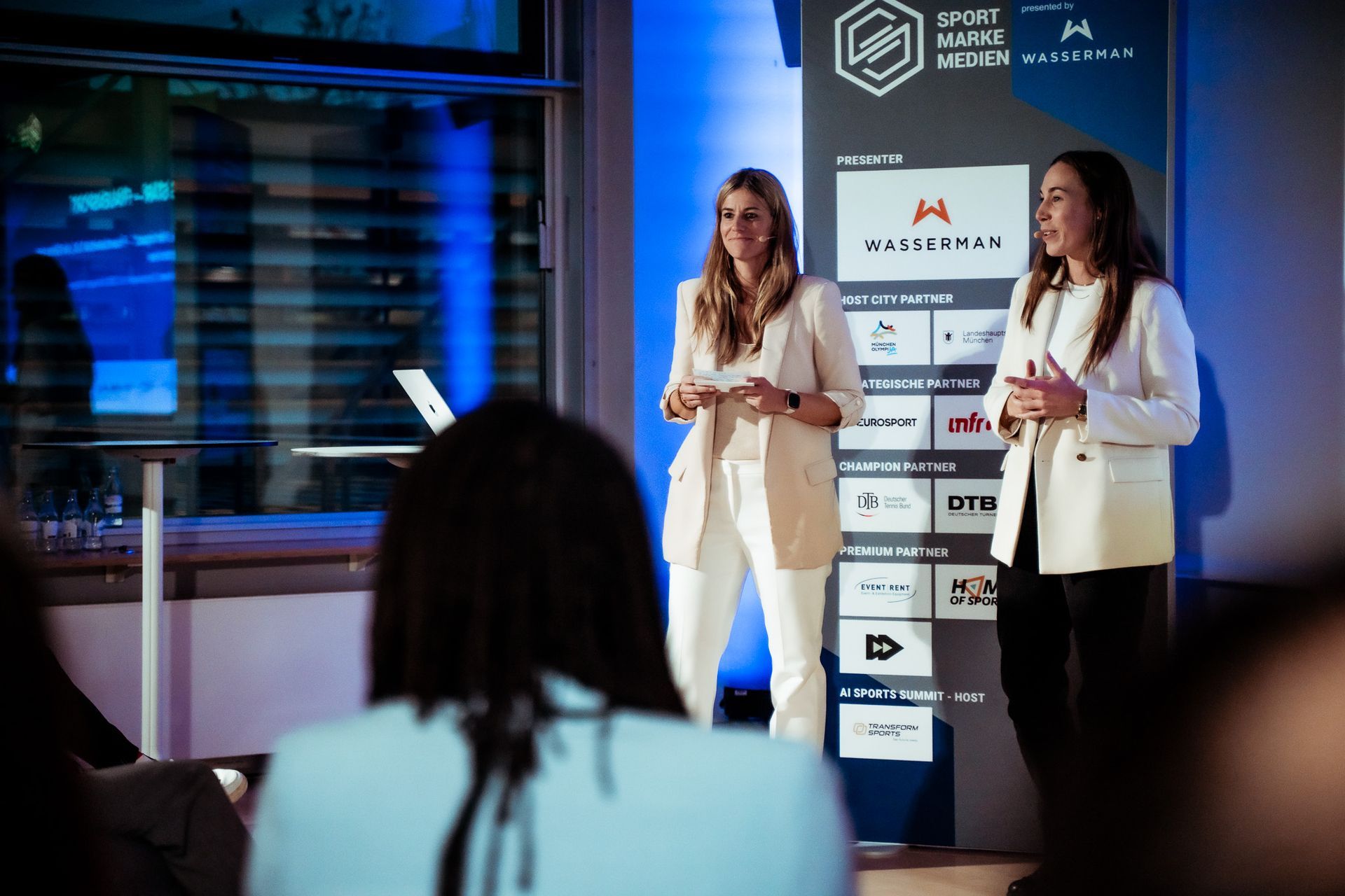 Two presenters on stage at a business event, standing before sponsor banners and audience silhouettes.