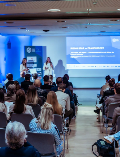 Audience watching two speakers present on a blue-lit conference stage with slides projected behind them