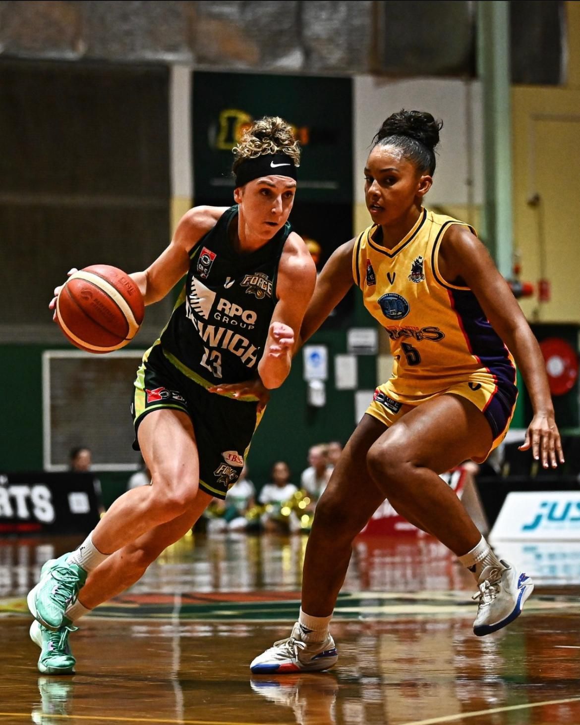 Two basketball players dribble on a polished indoor court during a game, one in black and one in yellow.