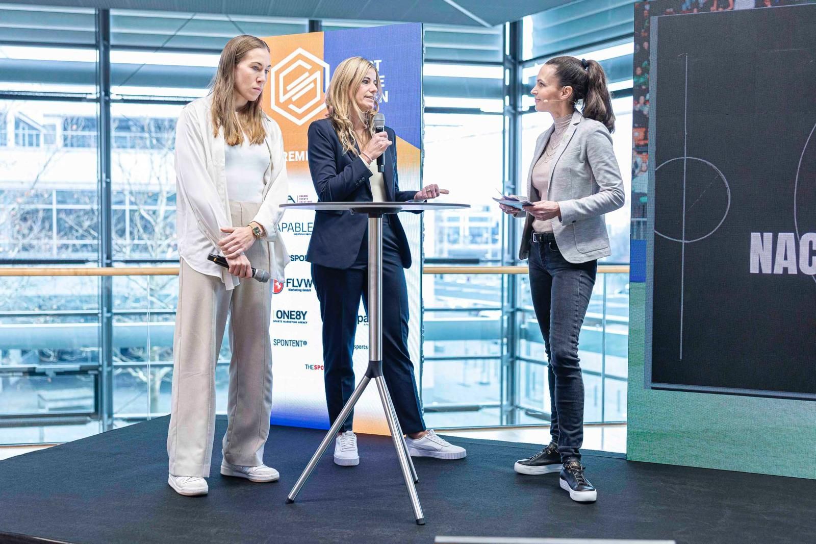 Three people stand around a tall table in a bright studio, presenting near a large screen.