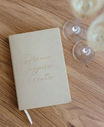 Cream notebook with gold text on a wooden table beside two wine glasses.