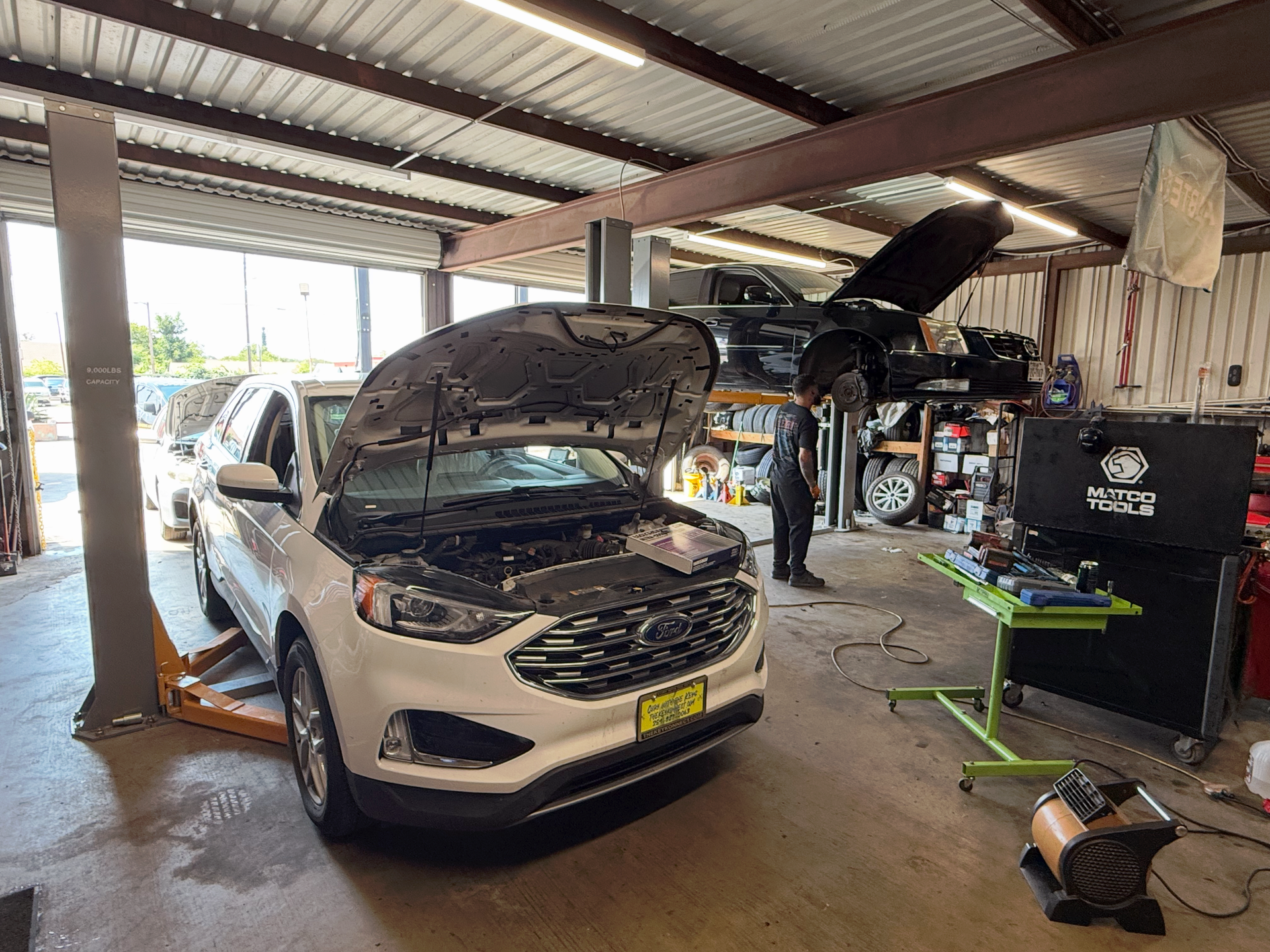 White SUV in auto repair shop, hood open. Mechanic near a black truck on lift. | Auto Masters Repair Shop