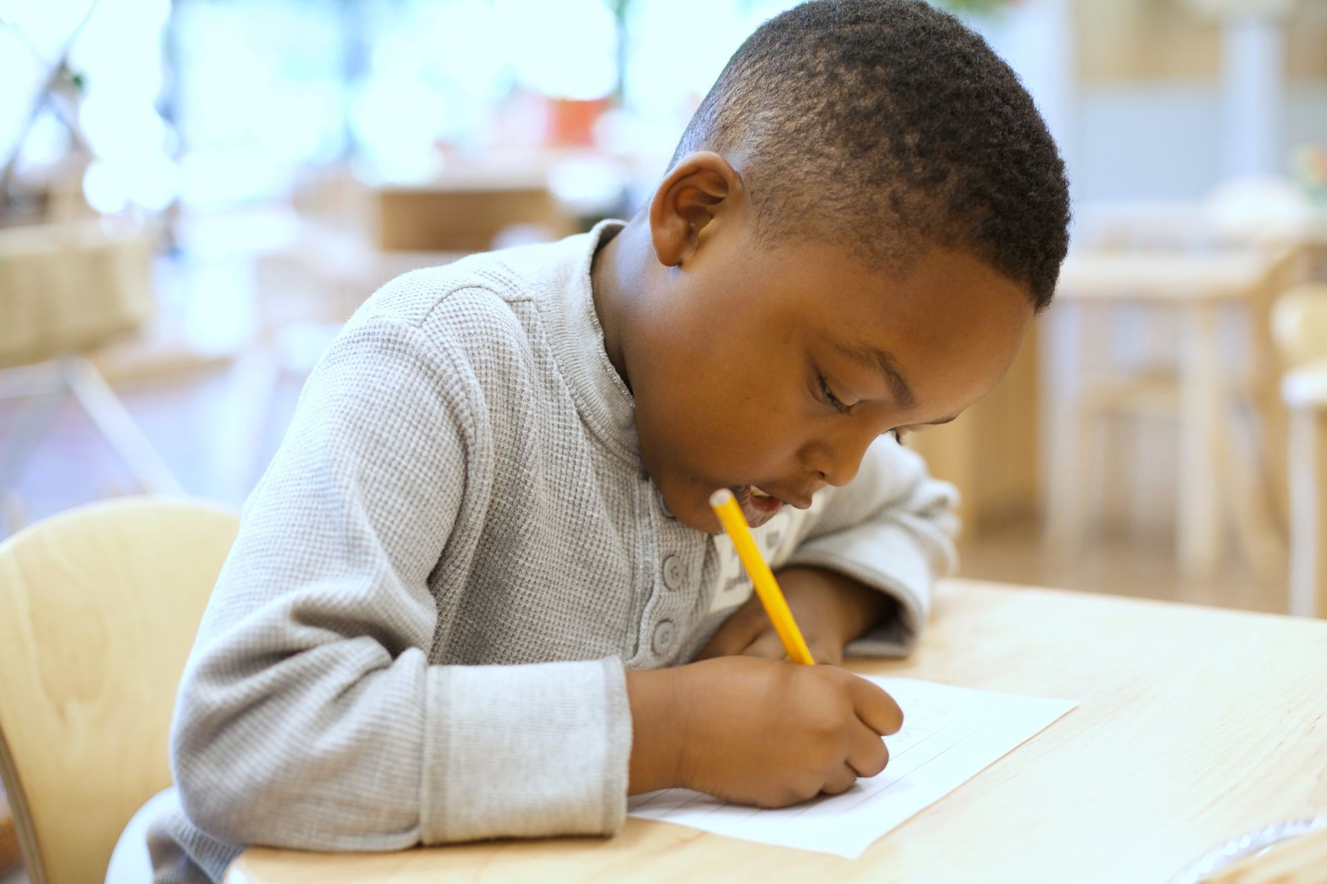 Child writing with a yellow pencil at a desk in a bright classroom