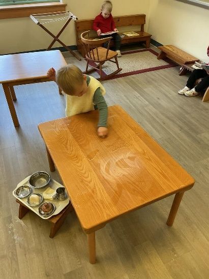Child interacting with a wooden table, small metal tray with cups nearby. Other children in background.
