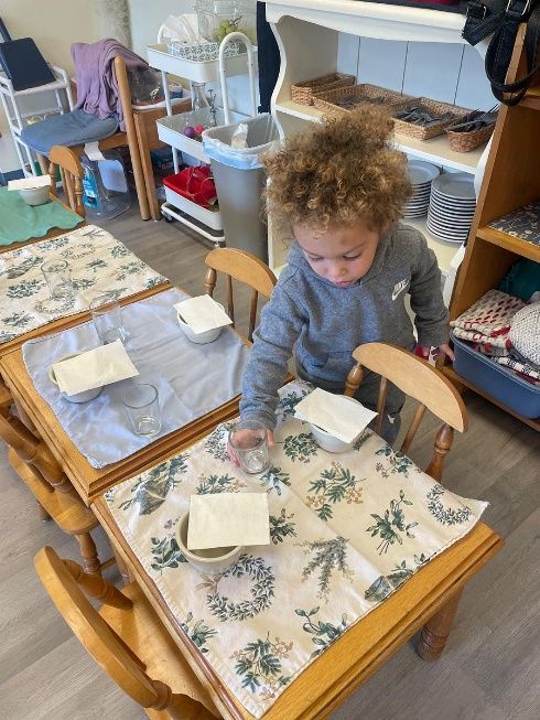 Child reaching toward a bowl on a table set with floral placemats in a well-lit room.