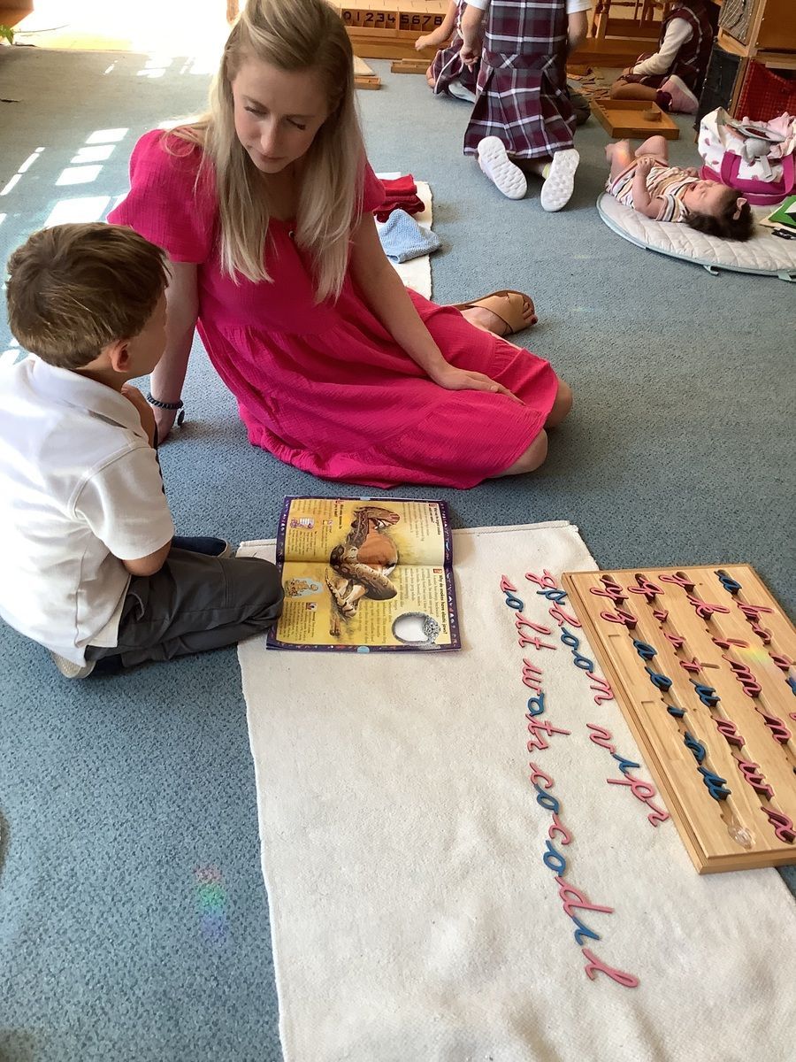 Children seated on a carpet, looking at a wooden moveable alphabet in a classroom.