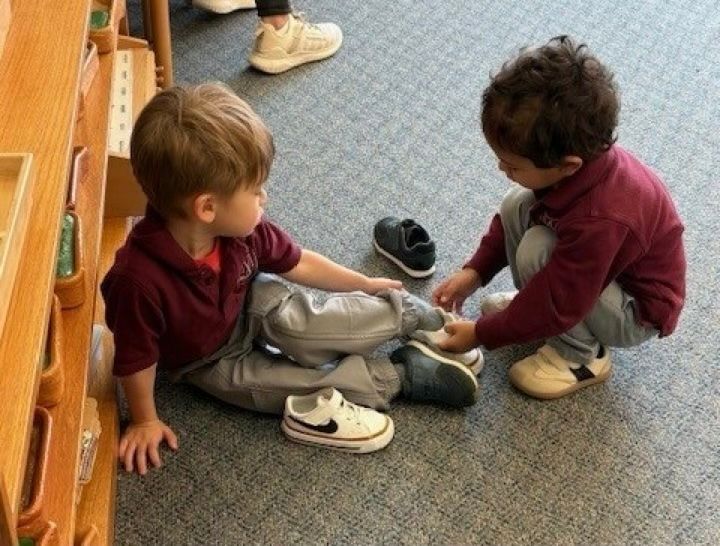 Two toddlers in maroon tops sit on a carpet, one helping the other with a shoe near a wooden shelf.