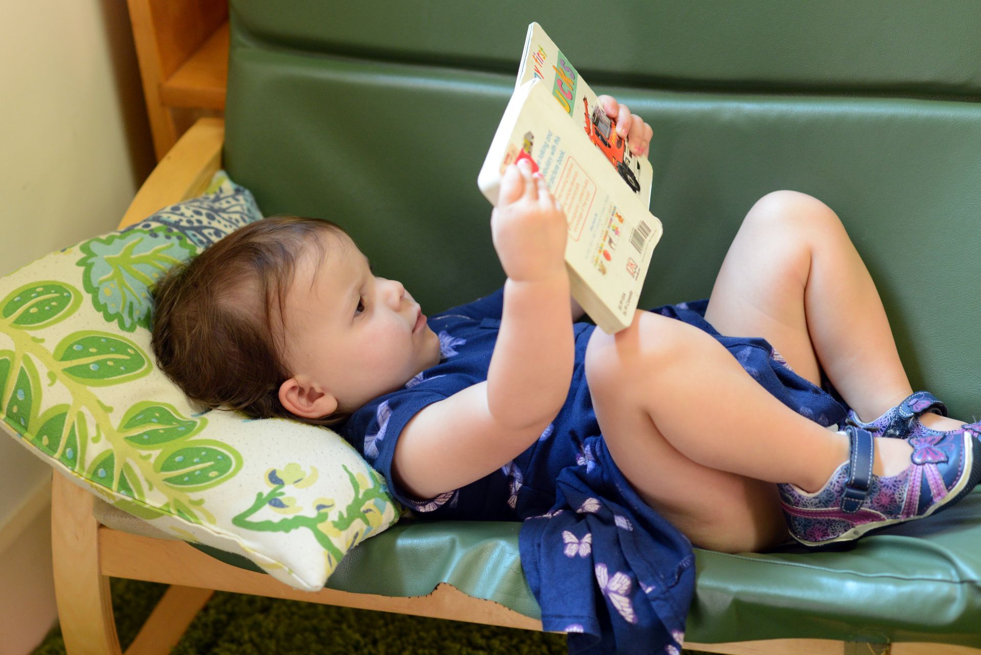 A toddler lying on a green chair, deeply engaged in reading a board book in a Montessori classroom.