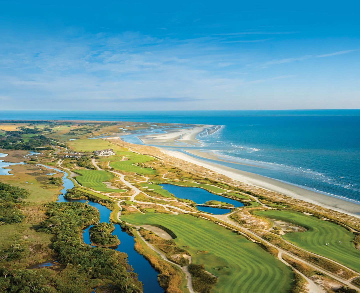 Aerial view of a coastal golf course with green fairways, blue ponds, sandy beach, and ocean