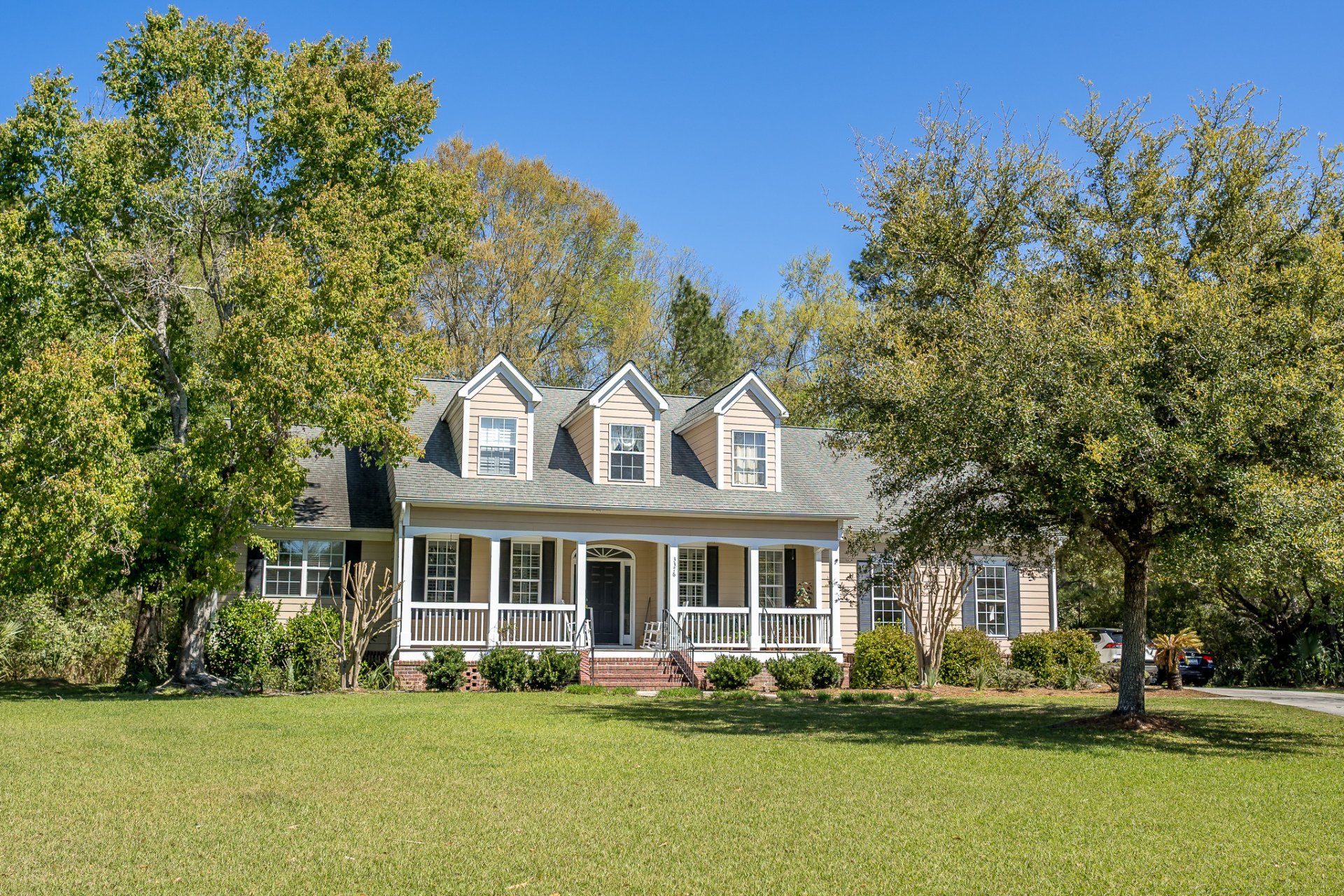 White farmhouse with dormer windows and a porch, surrounded by green lawn and large trees