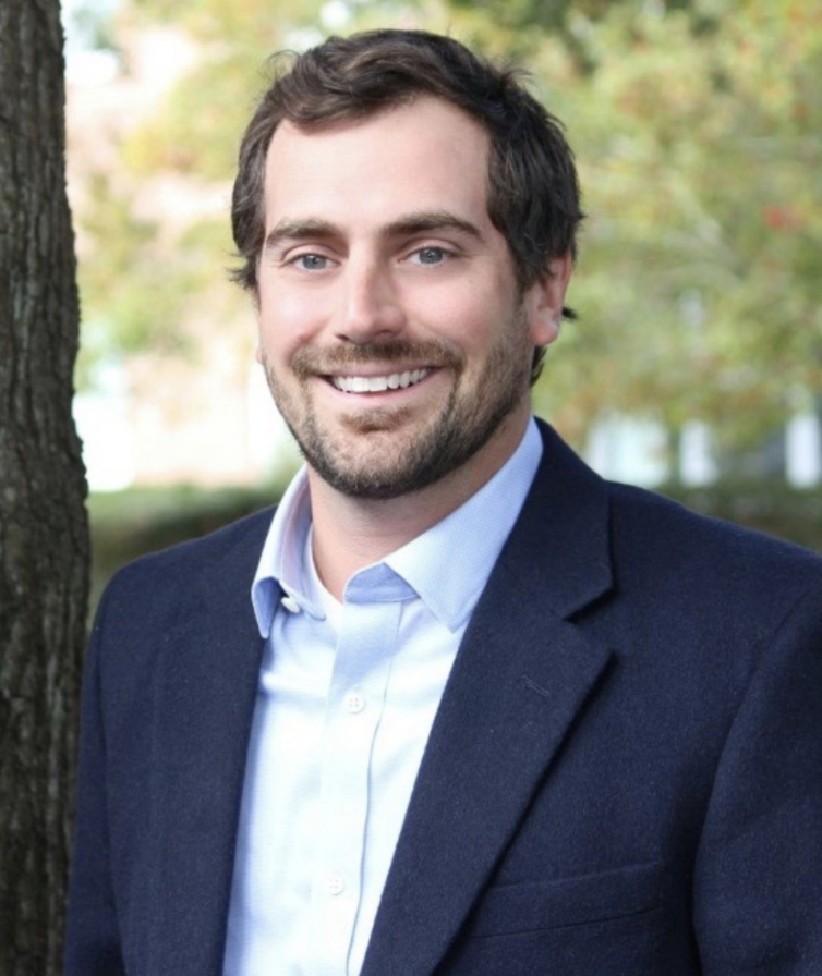 Smiling man in a navy blazer and light blue shirt outdoors with blurred trees in the background
