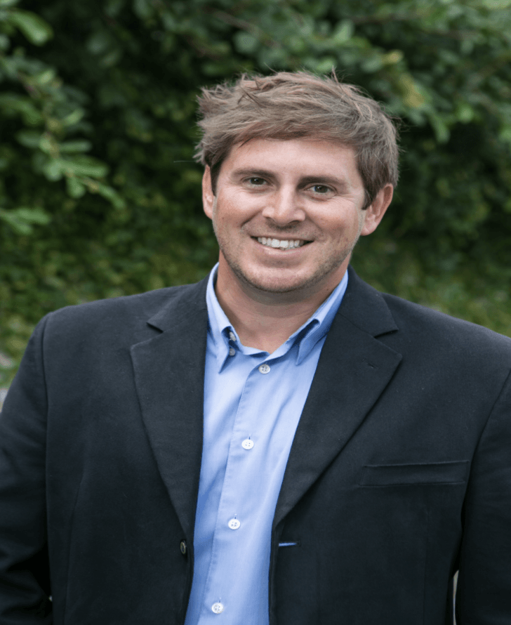 Man in a dark blazer and blue shirt smiling outdoors with green foliage behind him