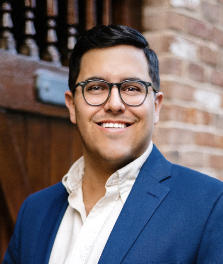 Smiling person in glasses and blue blazer standing against a brick wall