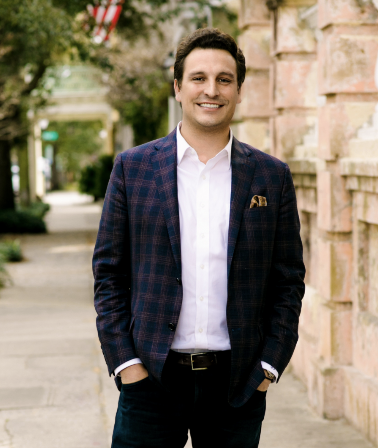 Man in a navy plaid blazer and white shirt smiling on a sidewalk beside a stone wall