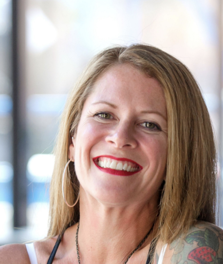 Smiling woman with red lipstick and a tattooed shoulder, seated indoors against a softly blurred background.