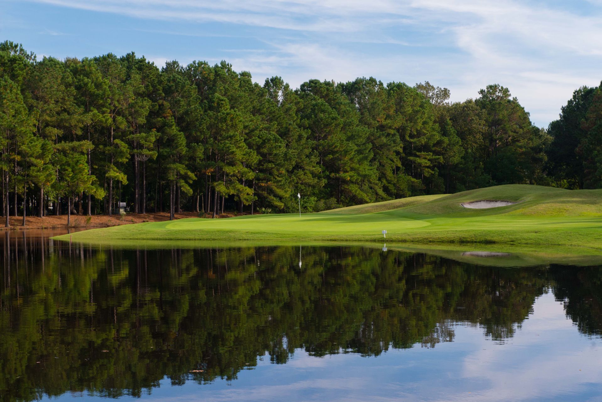 Golf course green by a calm pond, with dense trees and blue sky reflected in the water