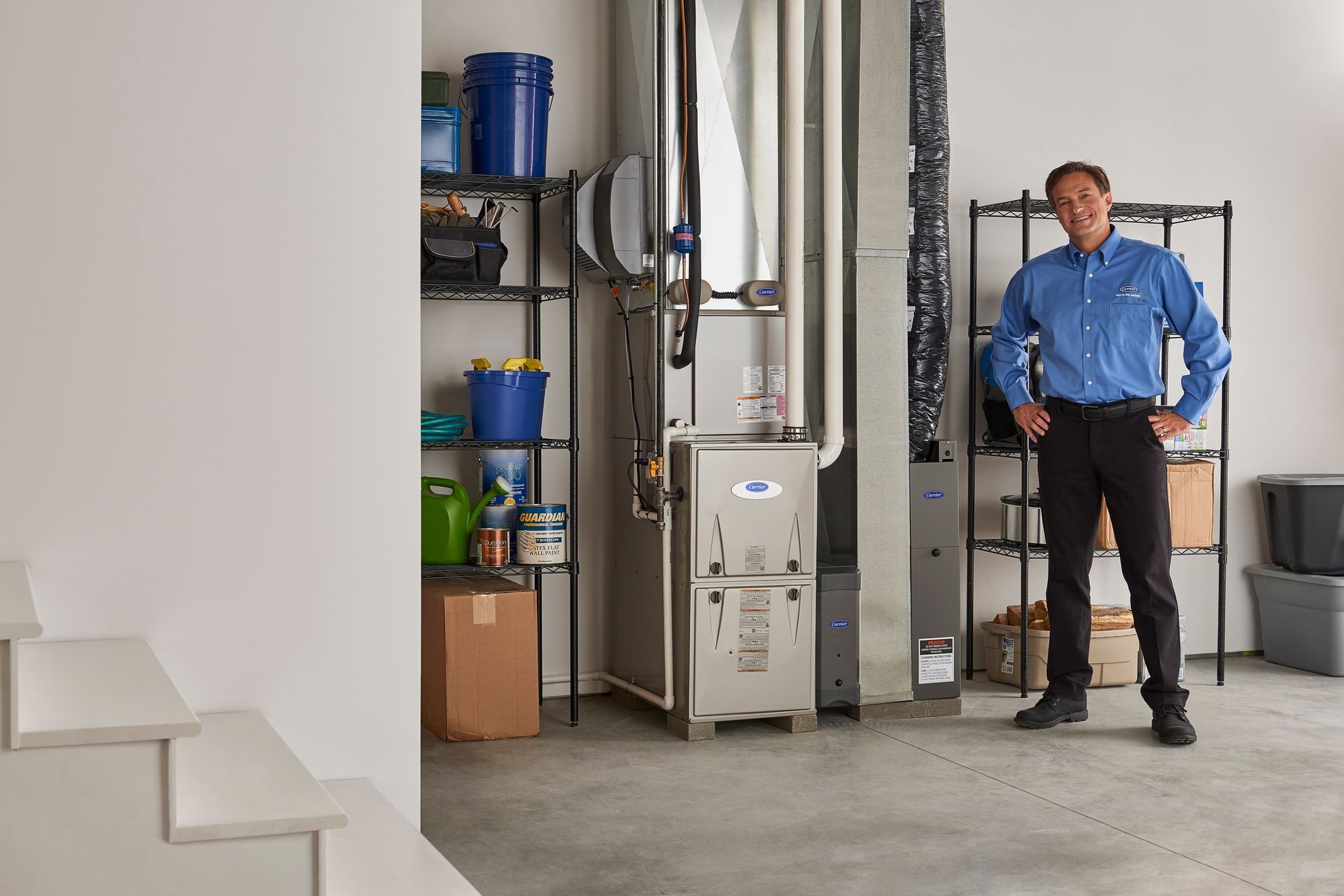 Man in blue shirt standing in a utility room beside HVAC equipment and shelves of supplies