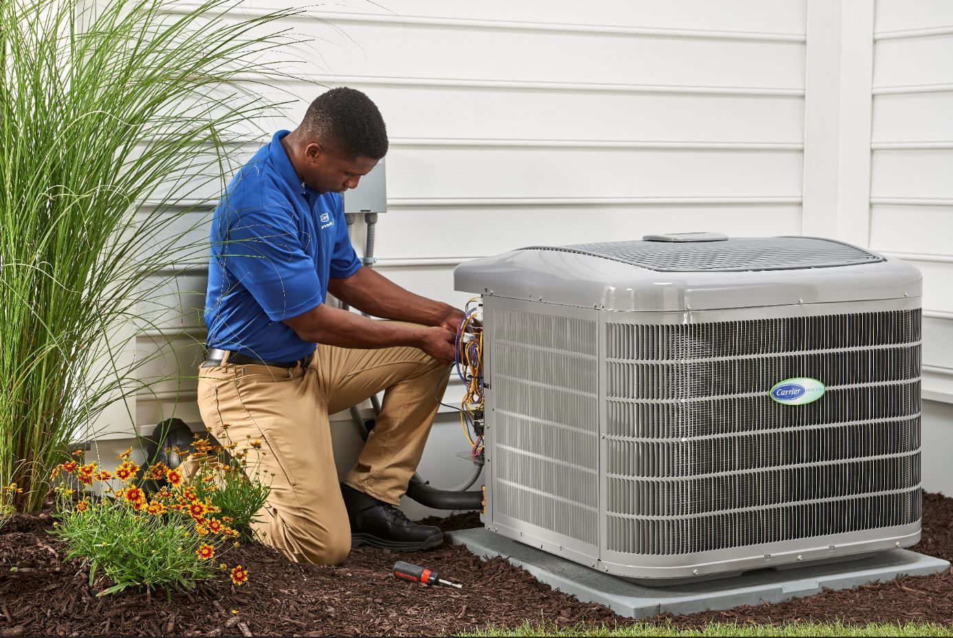 A service technician in a blue uniform kneels outside, repairing an HVAC unit next to a white house with landscaping.