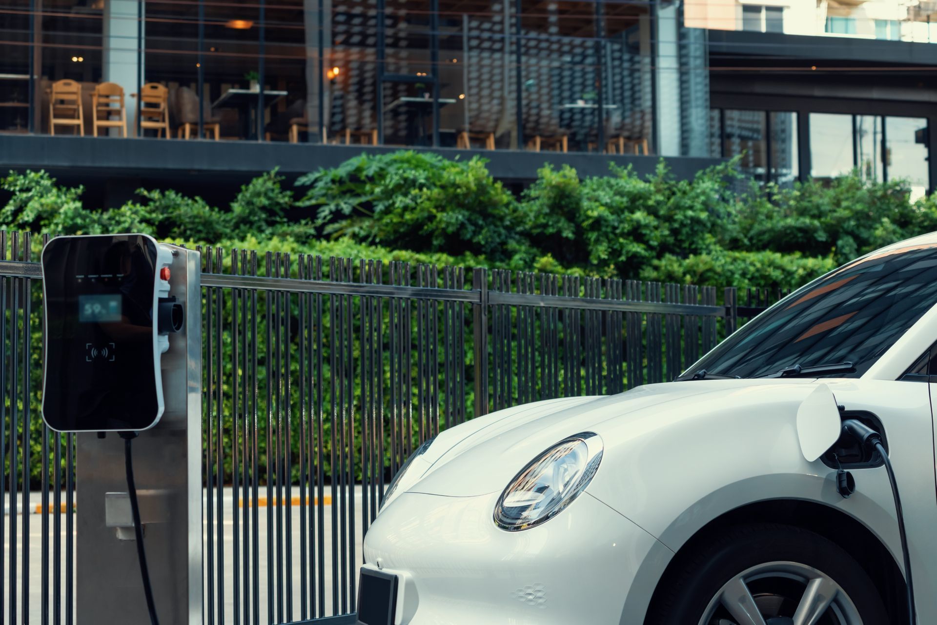 Electric car charging beside a modern building, with greenery and a fence in the background