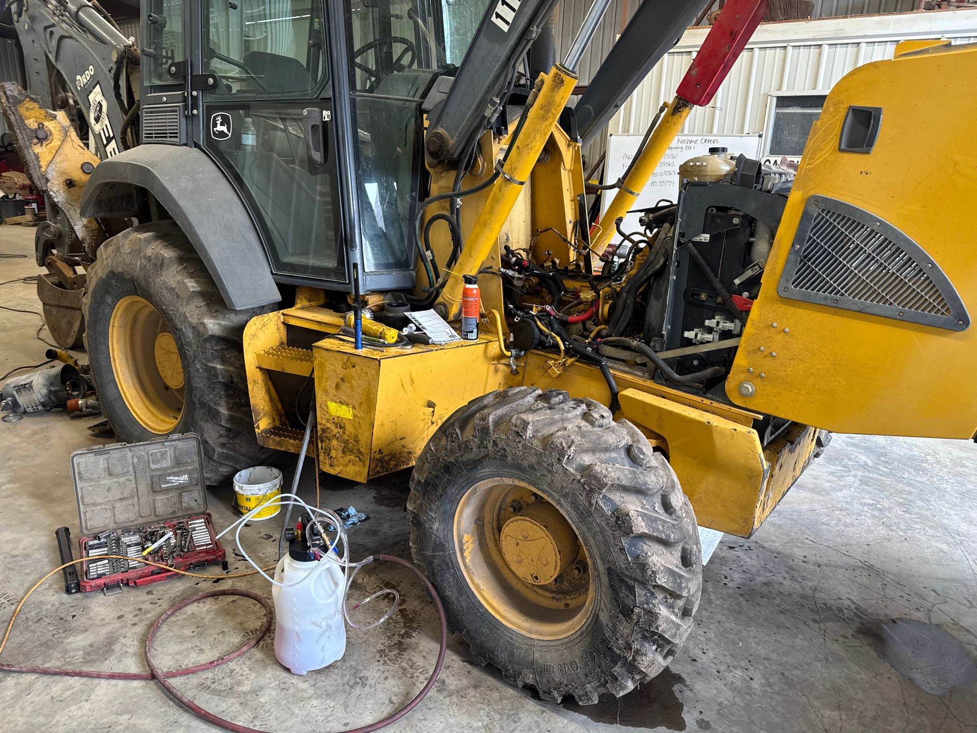 Yellow backhoe loader in a workshop, with front wheel removed and tools on the floor.