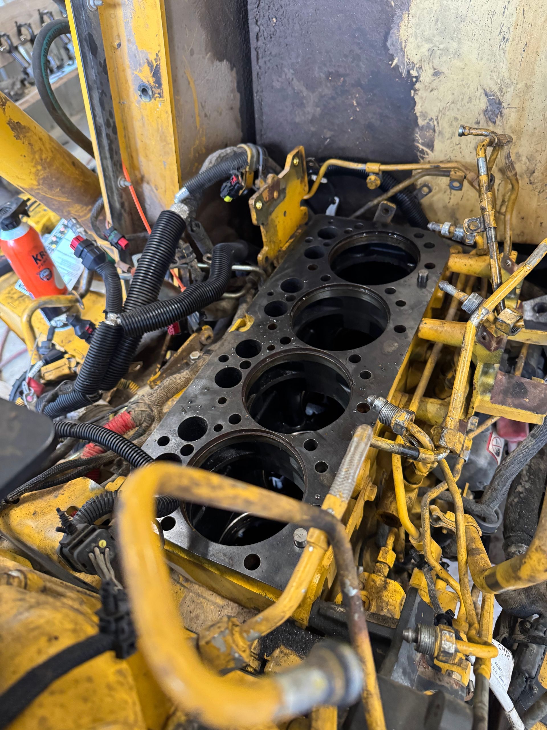 Engine block in a yellow workshop, with cylinder bores exposed and hoses and tools around it.