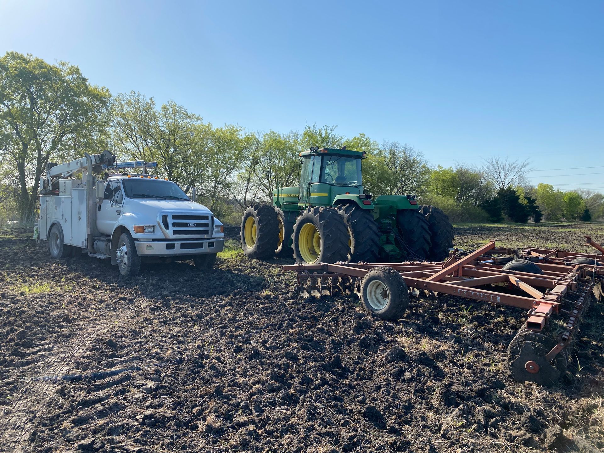 White utility truck and green tractor in a muddy field beside farm equipment under a clear blue sky