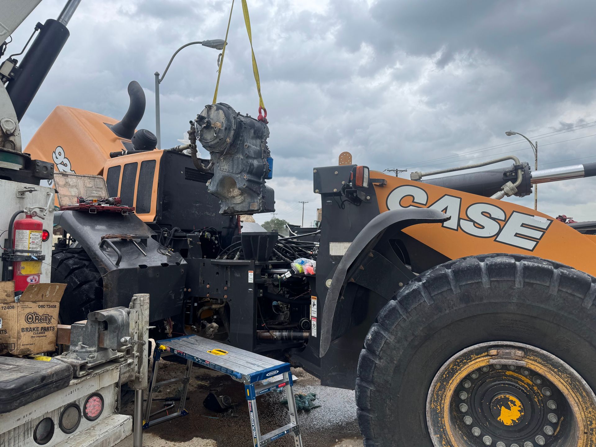 A CASE loader lifts a large engine part with a chain amid heavy equipment under cloudy skies.