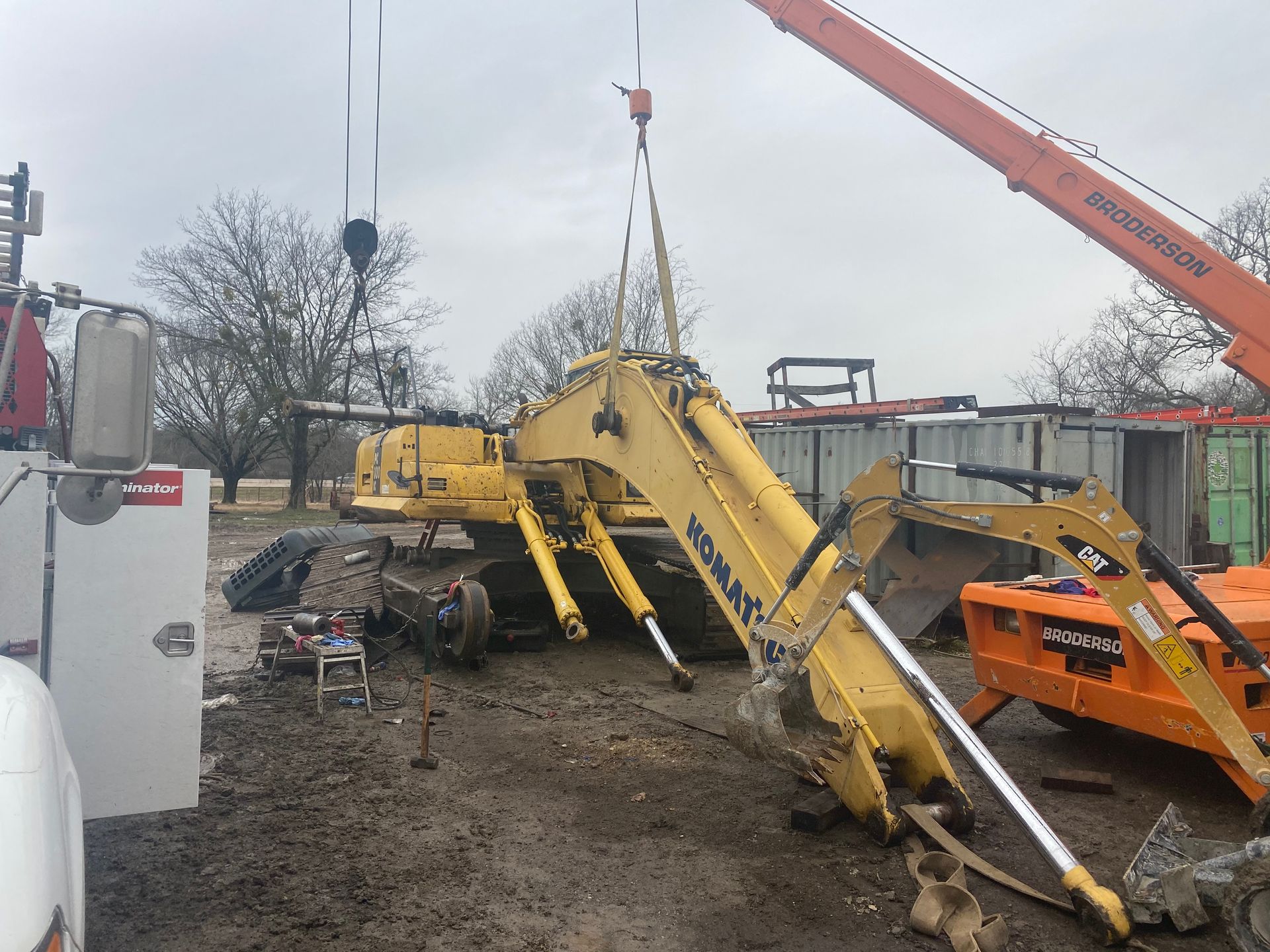 Yellow construction machinery being lifted by cranes in a muddy industrial yard