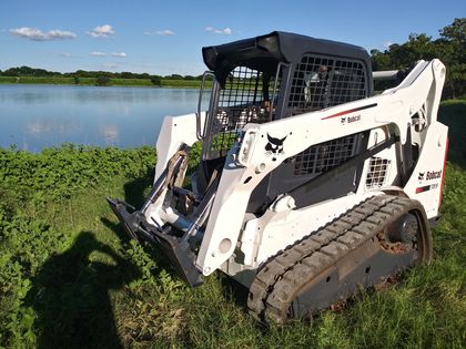 White skid-steer loader on muddy grass beside a lake under a blue sky