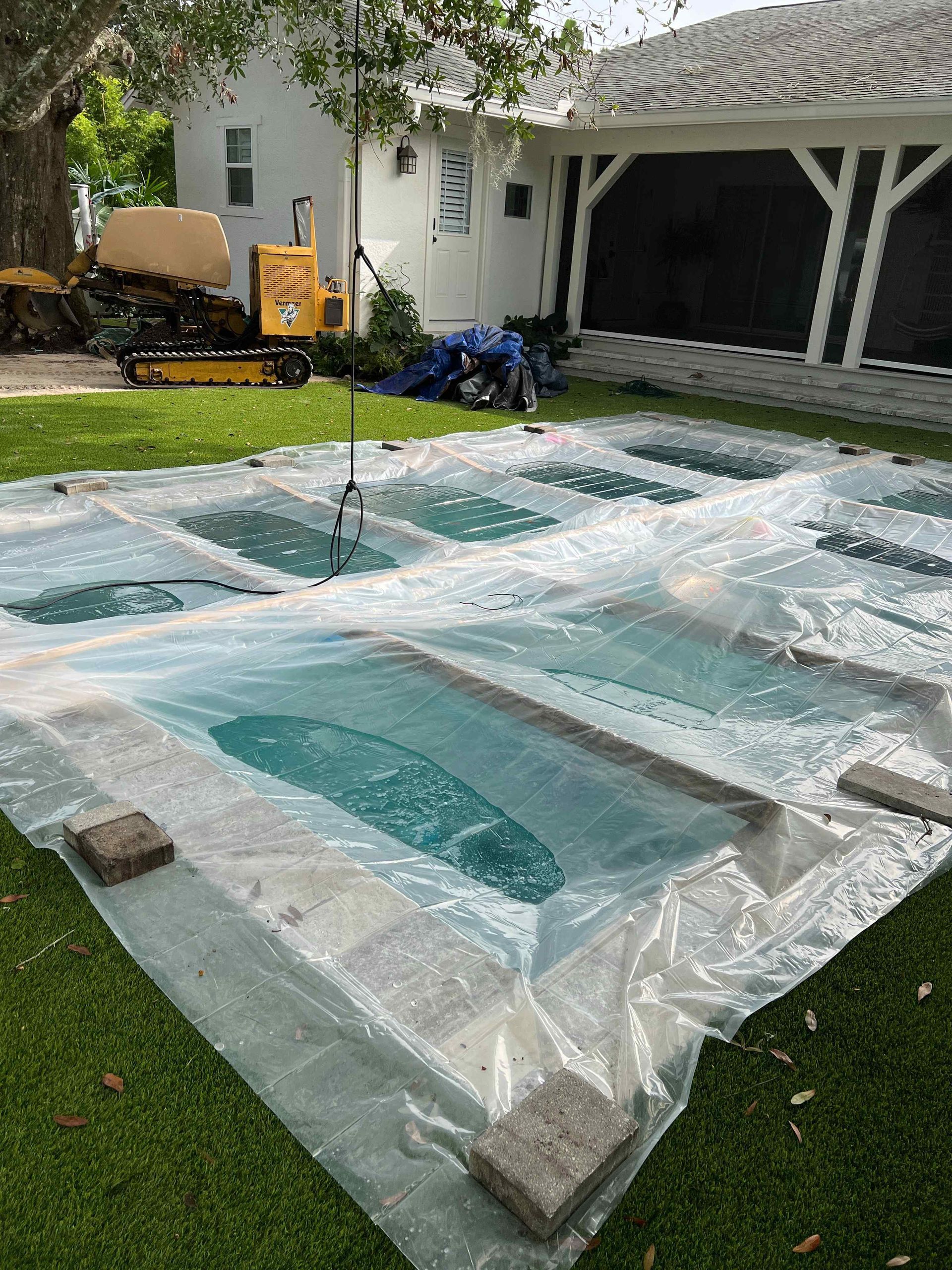 Rectangular water features covered in plastic on a green lawn, near a white building and garage.